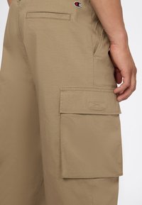 Beige cargoshorts met grote zijzak en het Champion-logo boven de achterzak, gedragen door een persoon met de hand rustend nabij de zak.
