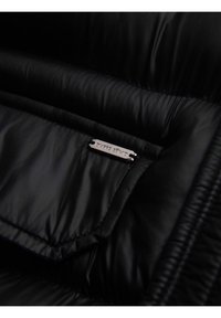 Primer plano de una chaqueta negra acolchada que muestra un bolsillo interior con una pequeña etiqueta metálica rectangular que dice "OKARE ESTAMO."