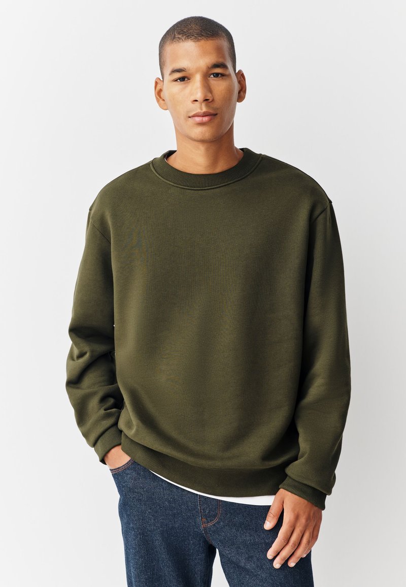 Olivgrön sweatshirt med rund halsringning, avslappnad passform, ribbade muddar och fåll, tillverkad av mjukt tyg, kombinerad med mörka jeans.