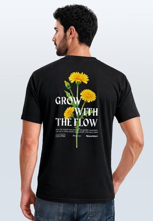 Man with short black hair wearing a black t-shirt with yellow dandelion flowers and text "Grow with the flow" on the back.