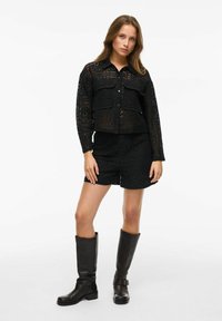 Crna čipkasta košulja s dva prednja džepa, uparen s crnim čipkastim šortsevima. Model nosi crne kožne čizme do koljena.
