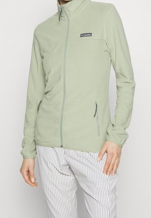 Personne portant une veste polaire Columbia vert clair avec des poches zippées et un pantalon blanc à rayures grises verticales.