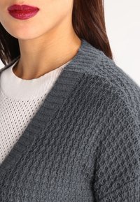 Grå texturerad kardiagan med ett stickat mönster, bärs över en vit mesh-top. Kardiagan har en bred krage och en avslappnad passform.