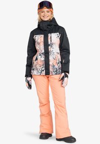 Black and floral patterned ski jacket with a zip front, paired with soft peach ski pants. Accessories include gloves and a beanie.