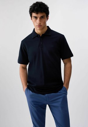 Giovane uomo in piedi con le mani nelle tasche, indossa una polo nera e pantaloni blu su uno sfondo chiaro e uniforme.