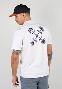 T-shirt en coton blanc avec un motif floral sombre au dos, à manches courtes et col rond. Porté avec un short gris et une casquette noire.
