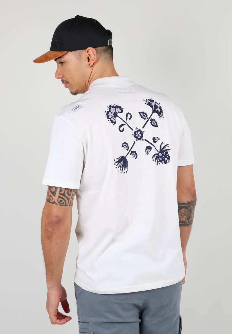 T-shirt en coton blanc avec un motif floral sombre au dos, à manches courtes et col rond. Porté avec un short gris et une casquette noire.