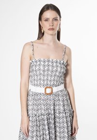 Vestido blanco con patrón floral negro, cuerpo fruncido y tirantes ajustables. Presenta un cinturón blanco con una hebilla cuadrada marrón.