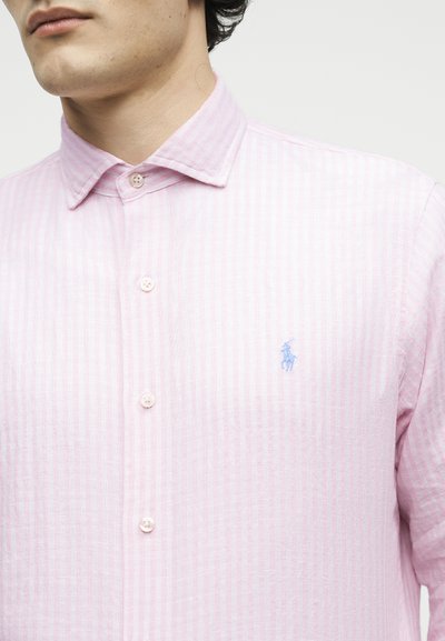 Άνδρας με ανοιχτό ροζ ριγέ πουκάμισο με κουμπιά και μικρό μπλε κεντημένο λογότυπο στο αριστερό στήθος, το γιακά κουμπωμένος.