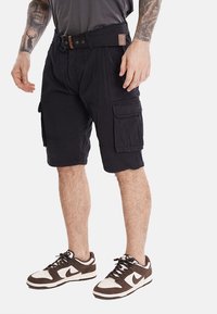 Pantaloni cargo neri realizzati in tessuto leggero, dotati di una cintura, numerose tasche e una vestibilità rilassata, abbinati a sneaker marroni e bianche.