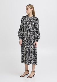 Vestido floral en blanco y negro con mangas largas, cintura ajustada y longitud midi. Presenta un cuerpo ajustado y una falda suelta y fluida.