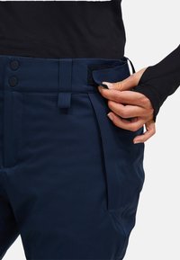 Mørkeblå bukser med en glat tekstur, udstyret med en knaplukning, velcrosnor og sidelommer, vist mens de justeres i taljen.