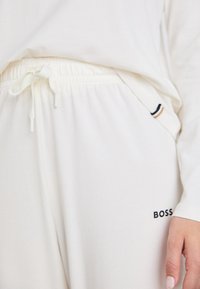 Joggeurs crème avec une taille élastique, un cordon de serrage, des poches latérales et le logo "BOSS" en noir au niveau de la hanche. Texture de tissu douce.