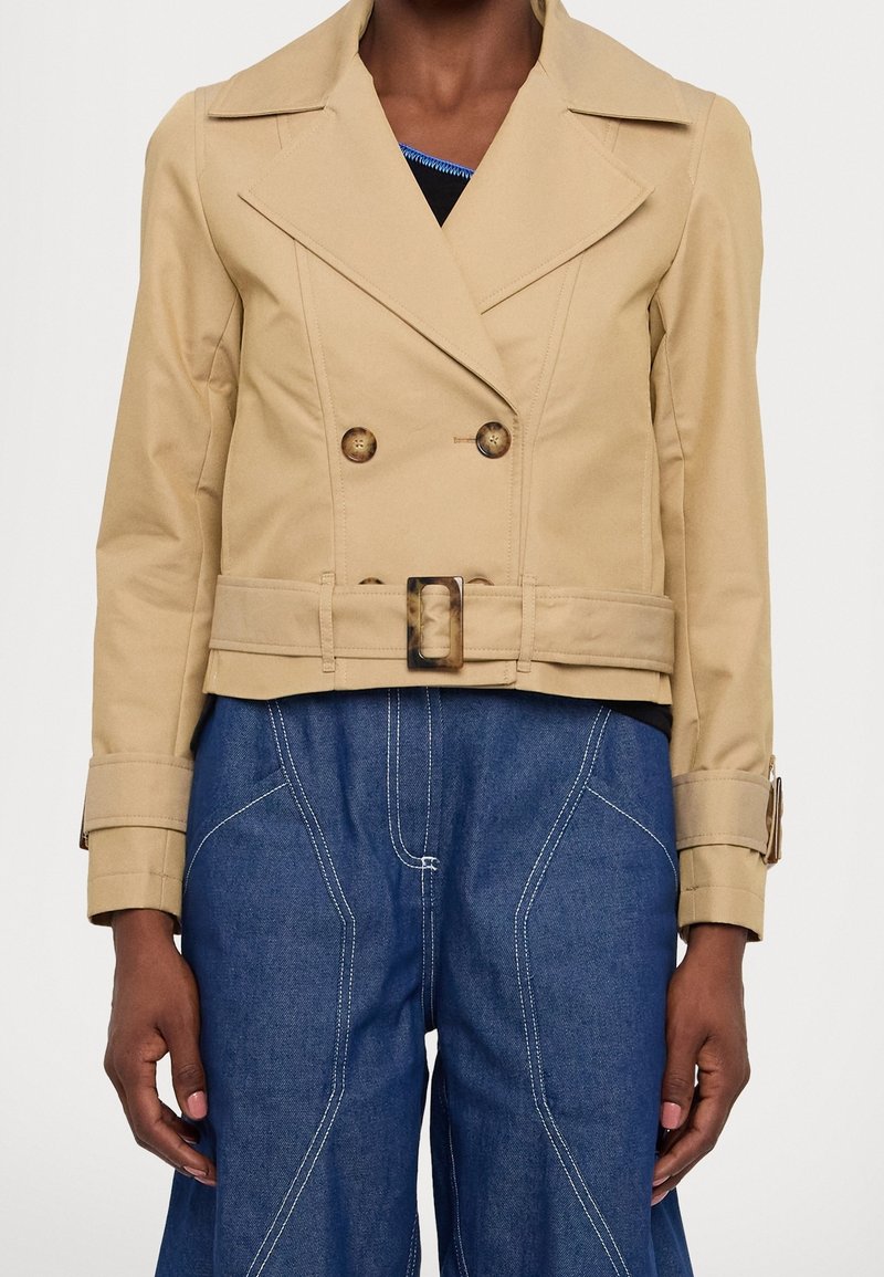 Beige dubbelbreasted cropped jasje met grote knopen, een ceintuur in de taille en brede revers. Draag het over denim met witte stikseldetails.