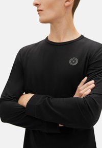 Zwarte long-sleeve shirt van zachte stof, met een ronde hals en een klein rond logo op de borst. Glad textuur.