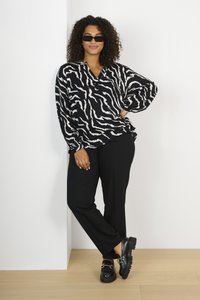 Blouse en imprimé zèbre noir et blanc à manches amples, associée à un pantalon noir et des chaussures noires imposantes. Le mannequin se tient de manière décontractée contre un mur.