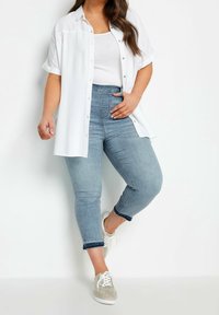 Camicia bianca a maniche corte con bottoni sopra una canottiera bianca, abbinata a jeans cropped azzurri e sneaker grigio chiaro. Look casual.