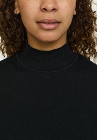 Črni rebrasti pulover s turtleneck ovratnikom, ki ima gladko teksturo in prilegajoč dizajn. Poudarjen je detajl ovratnika.