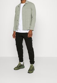 Ljusgrön knappjacka med två framfickor över en vit t-shirt, tillsammans med svarta cargobyxor och olivgröna sneakers.