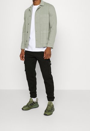 Světlé zelené zapínací sako s dvěma předními kapsami přes bílé tričko, zkombinované s černými cargo kalhotami a olivově zelenými teniskami.