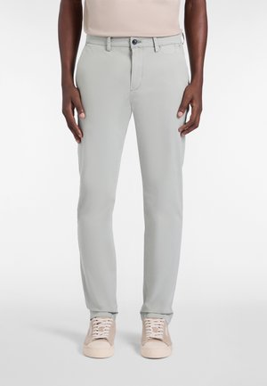 Man draagt lichtgrijze slim-fit broek en beige sneakers, staand tegen een effen witte achtergrond.