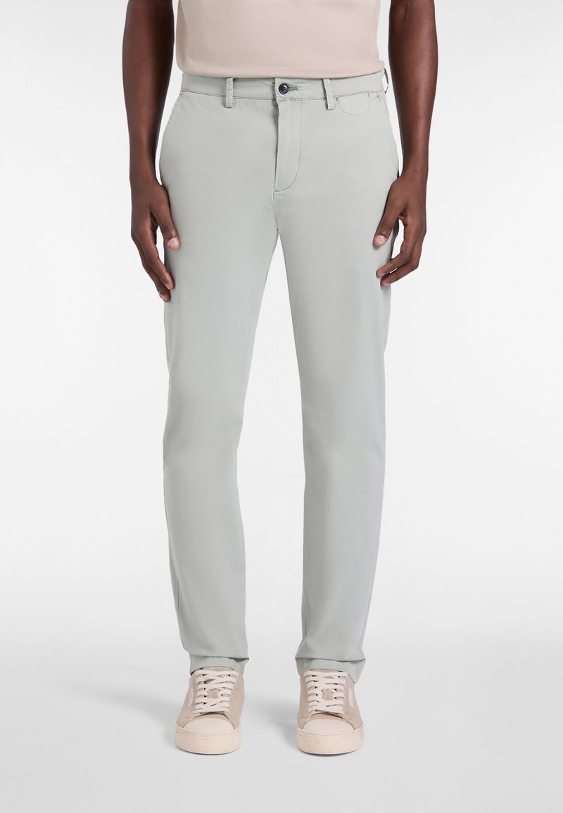 Mand iført lysegrå slim-fit bukser og beige sneakers, stående mod en ensfarvet hvid baggrund.