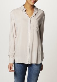 Vaaleanbeige, pitkähihainen paita, joka on valmistettu sileästä kankaasta. Siinä on napillinen edusta, kaulus ja rintatasku. Paidassa on kevyt laskeutuvuus ja ommelyksityiskohtia.