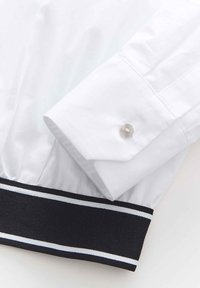 Manche d'une chemise blanche habillée avec une manchette à bouton unique reposant sur une bande en tissu noir avec deux fines rayures blanches.