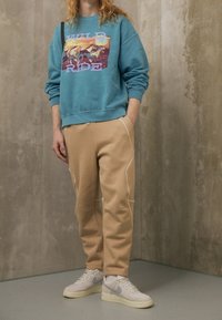 Tyrkysová mikina s nápisem "WILD RIDE" doplněná béžovými tepláky s bílými pruhy. Bílá obuv dokončuje celý outfit.