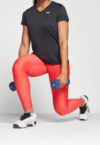 Svart kortärmad träningsskjorta, röda leggings, vit och svart sneakers, håller en blå hantel. Slät, åtsittande tyg, V-halsdesign.