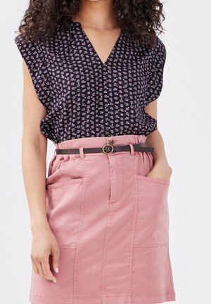 Woman wearing black floral sleeveless blouse tucked into high-waisted pink skirt with front pockets and a thin brown belt.