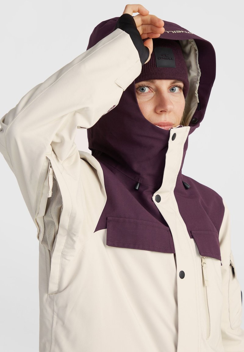 Kurtka narciarska w kolorze kremowym z głęboko fioletowym kapturem i akcentami, zapinanymi kieszeniami oraz regulowanymi mankietami zapewniającymi pewne dopasowanie.