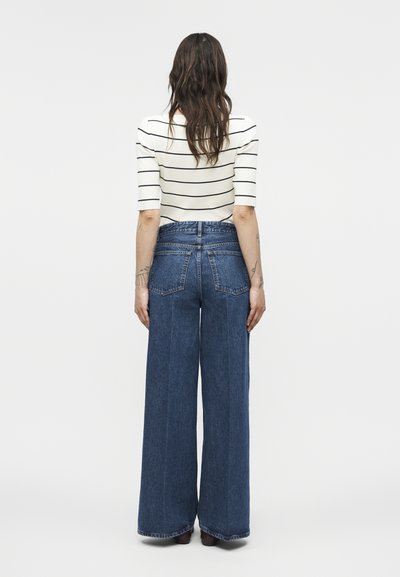 Žena s dlhými vlasmi, oblečená v bielom prúžkovanom topu a širokých modrých džínsach s vysokým pásom, stojaca otočená chrbtom na jednoduchom pozadí.