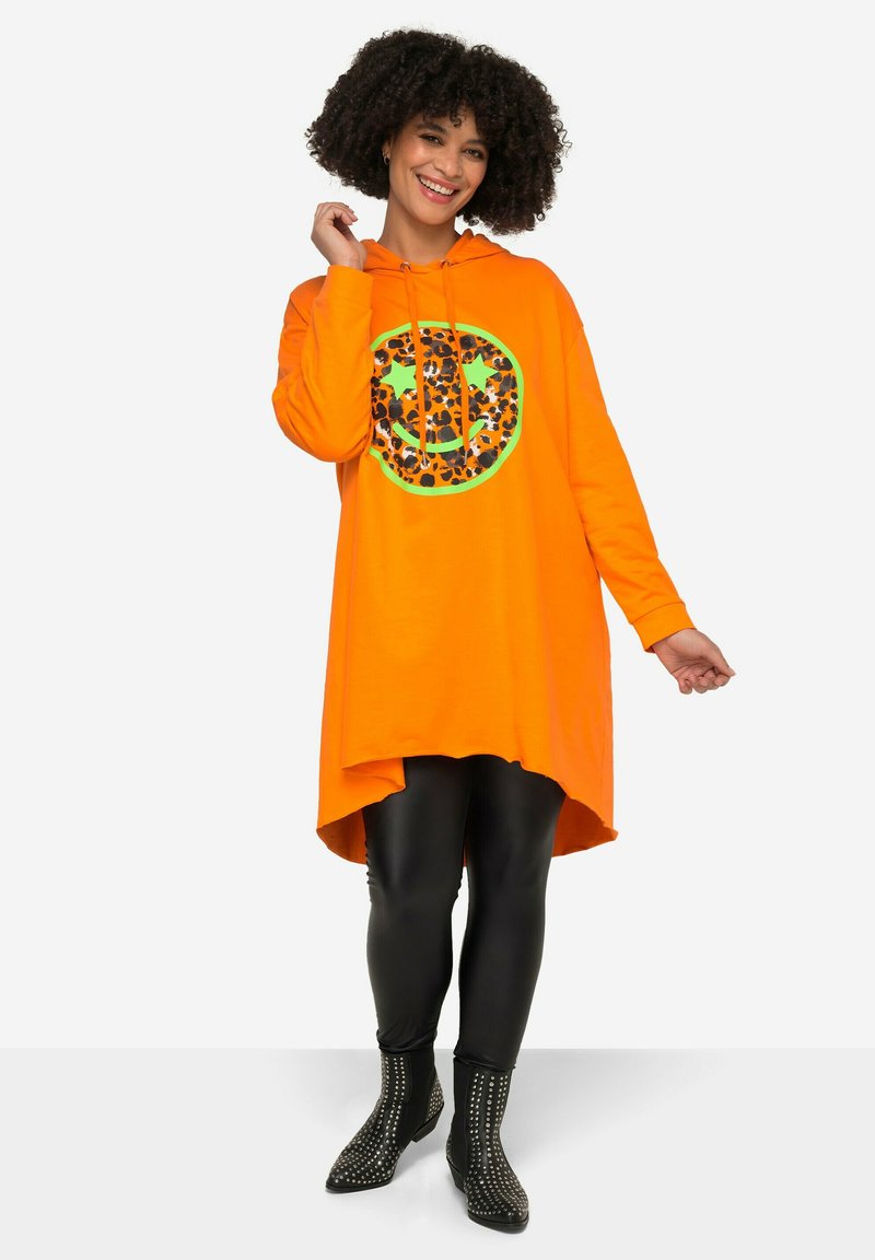 Vestito felpa arancione con grafica di un sorriso a motivo leopardato, maniche lunghe e orlo asimmetrico. Indossato con leggings neri e stivali alla caviglia con borchie.