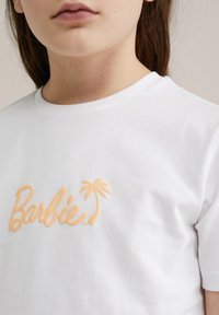 Camiseta blanca de algodón con logo bordado de "Barbie" en durazno y un acento de palmera. Cuello redondo y mangas cortas.