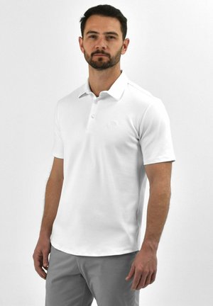 Man met donker haar en baard, gekleed in een wit poloshirt met korte mouwen en grijze broek, poserend tegen een effen witte achtergrond.