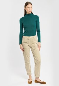 Maglione a collo alto a coste color teal, pantaloni chino beige e ballerine in suede marrone. L'outfit presenta un design attillato e polsini arrotolati sui pantaloni.