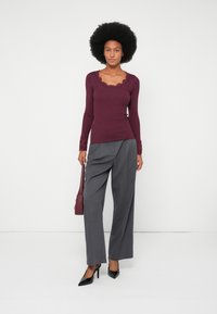Burgundy top s dlhým rukávom s volánovým výstrihom a čipkovými detailami, v kombinácii s vysokými šedými nohavicami a čiernymi špicatými lodičkami.