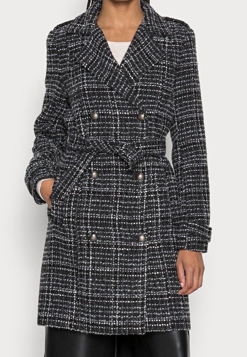 Cappotto doppiopetto a quadri bianco e nero con vita con cintura, ampi risvolti e dettagli con bottoni argentati. Realizzato in tessuto strutturato.