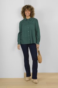 Green button-up shirt with a collar, tiered design, and long sleeves, paired with dark jeans and beige shoes, holding a tan handbag.