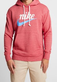 Man i röd Nike-hoodie med vita dragsnören och blå swoosh-logga, matchat med beiga byxor mot vit bakgrund.