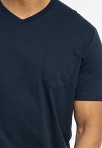 T-shirt in cotone blu navy con scollo a V e una piccola tasca sul petto. Maniche corte e vestibilità rilassata. Texture liscia con dettagli di cucitura minimi.