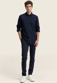 Laivastonsininen pitkähihainen nappipaita, yhdistetty tummiin housuihin ja valkoisiin tennareihin; kangas näyttää sileältä ja istuvalta.