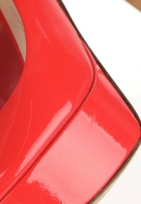 Gros plan d'une chaussure à talon haut en cuir verni rouge montrant une finition brillante et un détail d'orteil carré sur fond blanc.