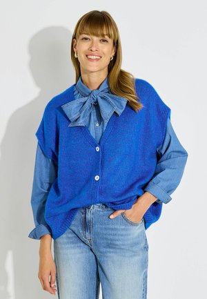 Mujer sonriente que lleva un chaleco de punto azul sobre una camisa vaquera con un gran lazo y vaqueros azul claro, de pie frente a un fondo blanco.