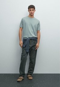 Ljusblå kortärmad t-shirt, mörkgrå lösa jeans, marinblå tröja knuten runt midjan, beige tofflor, stående mot en enfärgad bakgrund.