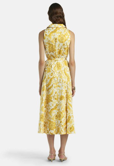 Kvinde iført hvid, ærmeløs midikjole med gult blomsterprint, stående med ryggen til, kombineret med lavhælede sandaler og armbånd.