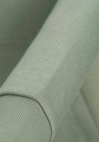 Világoszöld textil, sima felülettel és bordázott díszítéssel. Közeli felvétel az ujjon, amely kiemeli a varrás részleteit és a szövet puhaságát.