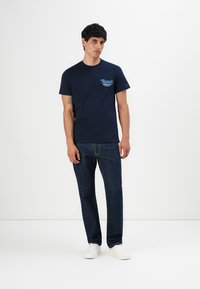 Homme portant un t-shirt graphique Tommy Hilfiger bleu marine, un jean bleu foncé et des baskets blanches, se tenant devant un fond blanc uni.