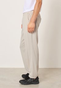 Šoninis vaizdas į asmenį, dėvintį šviesiai smėlio spalvos rupias kelnes, baltą marškinėlį ir juodus sportinius batelius, stovintį ant lygios grindų dangos.